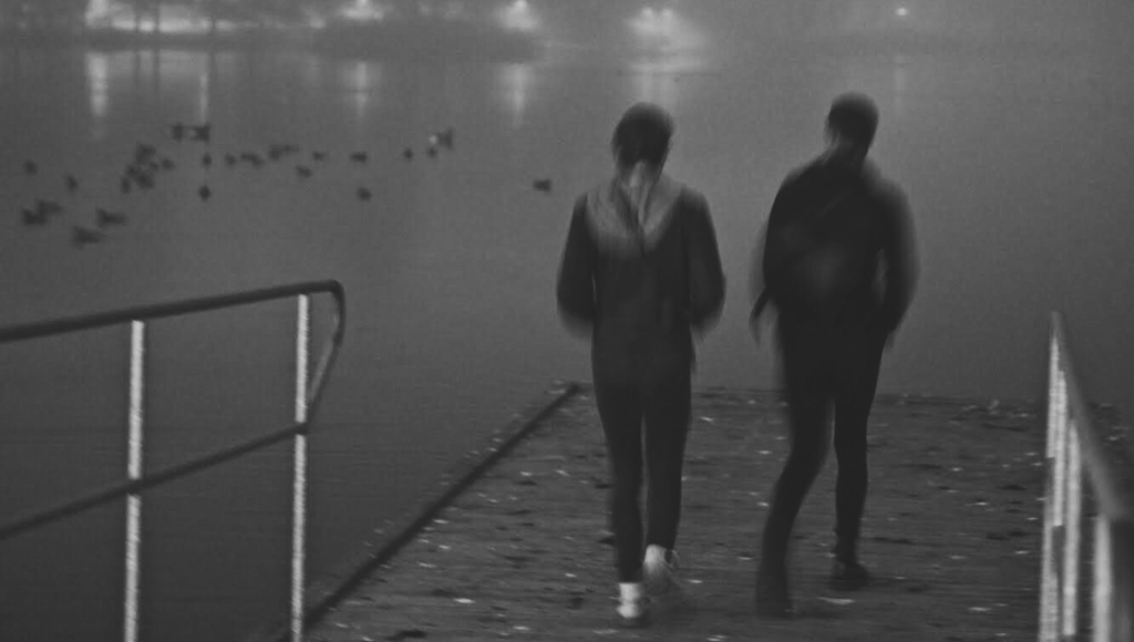 Kaksi nuorta seisoo laiturilla ja katsoo merelle päin, mustavalkoinen kuva.