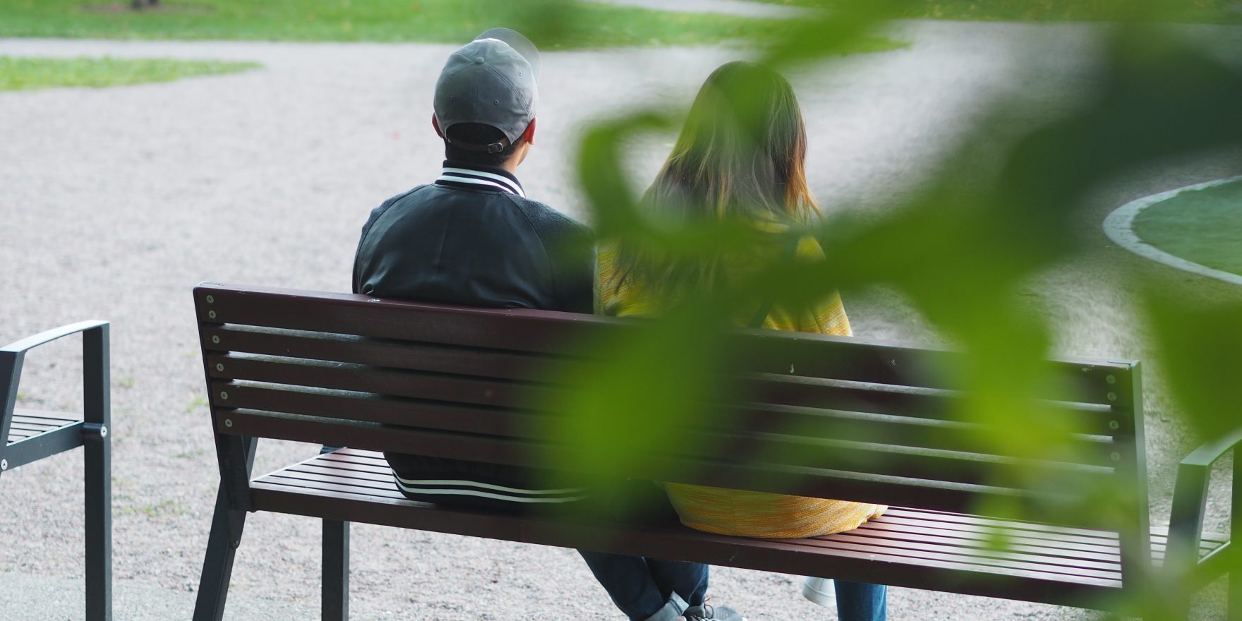 Kaksi ihmistä istuu leikkipuiston kentällä, katsoen poispäin kamerasta kohti leikkipuistoa.